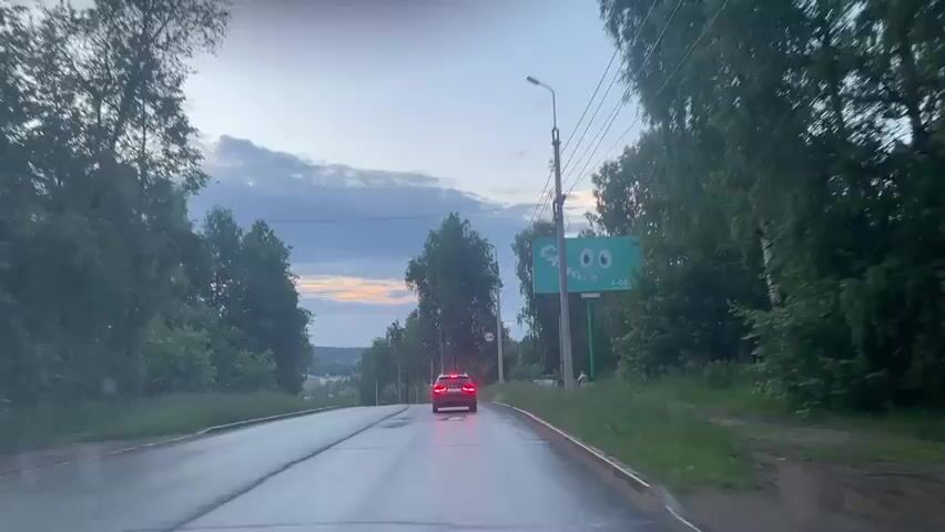 The video shows a rear-view perspective from inside a vehicle driving on a wet asphalt road during what appears to be dusk or twilight. The sky is cloudy with hints of orange and yellow from the setting sun. Trees line both sides of the road, with lush green grass and foliage. Ahead, a red car is visible, its brake lights illuminated. To the right of the road, a large billboard with a cartoonish face and Russian text is present, along with utility poles and wires. The road surface is reflective, indicating recent rain. The overall environment suggests a rural or suburban setting.