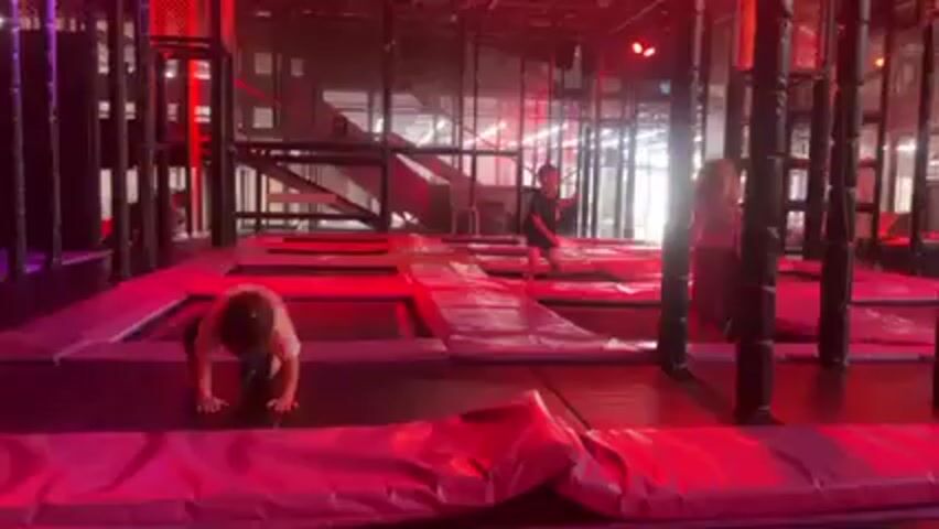 The video shows an indoor trampoline park with multiple children playing. The lighting is a combination of red and purple, creating a vibrant atmosphere. One young child is jumping on a trampoline in the foreground, wearing a light pink and white shirt with black shorts and white sneakers. Two other children are seen further away, also jumping on trampolines. The trampoline park has padded borders and columns, with a multi-level climbing structure visible in the background.
