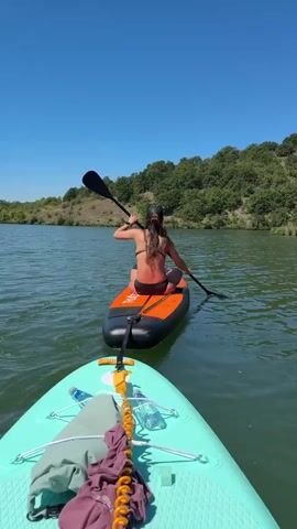 Two paddleboards are on a body of water. The closer paddleboard is light blue and has gear strapped to its front, including a green bag, a maroon towel, and a coiled yellow cord. The further paddleboard is orange and black, with a woman sitting on it. She is wearing a black bikini and a baseball cap, and she is holding a black paddle and appears to be paddling. The water is calm and reflects the clear blue sky. The background shows a shoreline covered in green trees.