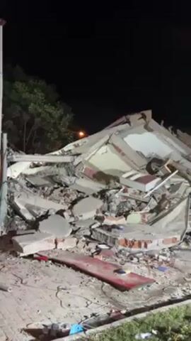 The video shows a demolished building at night. The building appears to be constructed of brick and concrete. The structure is in a state of complete collapse, with debris scattered across the surrounding area. There are some street lights providing illumination and a few trees visible in the background. The environment seems to be a built-up area, likely a city or town.