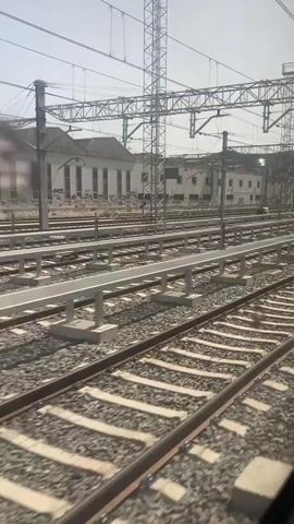 A first-person perspective video shows a view from a moving train. Multiple train tracks stretch out in parallel, leading towards a cluster of white buildings in the background. Overhead, a complex network of catenary wires and supporting poles is visible, indicating an electrified railway. The ground beneath the tracks is covered in ballast. The sky is bright and clear, suggesting daytime. There are no people clearly visible. The train appears to be in a station or railyard, possibly preparing to depart or having just arrived.