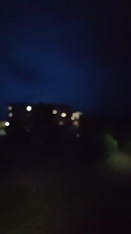 The video shows a dark nighttime scene with a building in the distance. The sky is a deep blue, with some clouds. The building appears to be illuminated by interior lights, with a few windows lit up. There are trees and other foliage in the foreground and around the building, which are mostly obscured by darkness. A faint light is visible on the right side of the frame, possibly from a vehicle or street lamp. The overall impression is of a quiet, nocturnal setting.