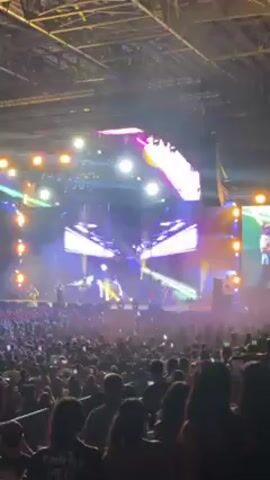 The video captures a concert scene indoors. A large crowd faces a brightly lit stage featuring a band and a large screen displaying animated graphics and lighting effects in various colors, including green, purple, yellow, and white. The crowd appears engaged, watching the performance. A disco ball hangs from the ceiling. The venue has high ceilings and is filled with sound and light equipment.