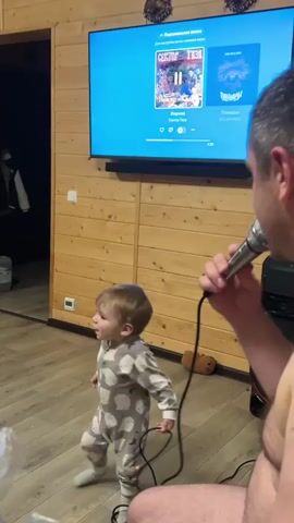 A man wearing no shirt sings into a microphone, standing indoors while his young child in a patterned, one-piece pajama suit stands in front of him. The child holds the microphone cord. In the background, a flat screen television displays the playing music. The room has wood paneled walls, and the screen and speaker bar are mounted to it. The light wood floor is shiny. An open doorway is visible in the background, with a dark interior and a white jacket hanging on the inside of the door. Animal head decorations are affixed to the wall above the television.