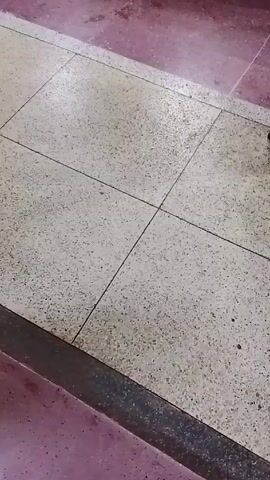 A person wearing a long garment and dark shoes walks across a tiled floor with a gray and white speckled pattern. The floor is bordered by a pinkish-red surface. The person's footsteps are captured as they move from right to left in the frame. The camera angle is a low shot, focusing on the lower part of the person's body and the floor. The scene appears to be an interior setting, likely a hallway or public space.