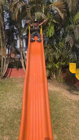 A young boy wearing a black cap, glasses, an olive green t-shirt, a quilted vest, dark gray pants, and black and white sneakers goes down an orange slide in a park. He yells "Yeah!" as he reaches the bottom. He then walks away from the slide, facing away from the camera. The park has palm trees, a brick wall, and a yellow bench in the background.  The grass is sparse and appears to be slightly brown in places. A white building with windows can be seen in the background as well.