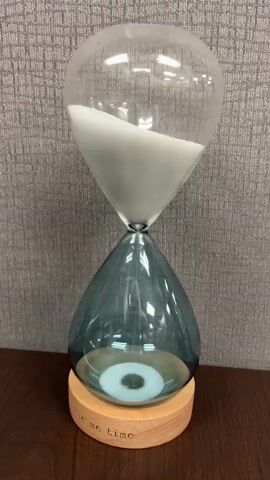A close-up shot shows an hourglass with clear glass and a teal glass bulb sitting on a wooden base. White sand is slowly falling from the top bulb to the bottom bulb. The hourglass is placed on a wooden surface with a gray textured background. The base of the hourglass has text written on it which says "me time".