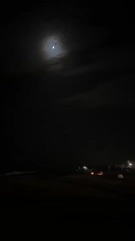 The video depicts a nighttime scene at Playa Del Sol-Villacana, Spain. The moon is visible in the sky, partially obscured by clouds, casting a dim light on the surroundings. The setting appears to be a beach or a coastal area, with the dark ocean visible on the left side of the frame.

On the shore, several tents are pitched, suggesting a camping or outdoor gathering. A bonfire is burning brightly, illuminating a group of people gathered around it. Their silhouettes are visible, and the fire casts a warm glow, creating a cozy and communal atmosphere. The presence of tents and the bonfire indicates that people are spending the night outdoors, possibly enjoying a camping trip or a beach party.

The time of day is clearly night due to the darkness and the illuminated moon. The weather seems calm, with no strong winds or rain apparent. The location is identifiable as Playa Del Sol-Villacana, Spain, a coastal resort area. The overall mood conveyed is one of relaxed enjoyment and camaraderie, with the warm fire and the presence of people creating a sense of community against the dark, vast backdrop of the night sky and sea. There is no visible text in the video.