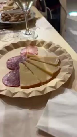 The video shows a wooden table with several plates of food and glasses of white wine. The plates contain salami, cheese, ham, and breadsticks. A woman in a white coat stands next to the table and takes a bite of something. Other people are standing in the background. There's a metal container and an aluminum utensil on the table. The table is decorated with the words "DESDE EL SER." The video appears to have been taken indoors, possibly at a bar or restaurant.