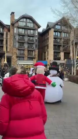 The video shows an outdoor scene in what appears to be a plaza or walkway in a town or resort area. Several people are walking around. The background includes large buildings with multiple stories and balconies. The architecture suggests a mountain resort or upscale town. In the foreground, there are three inflatable Christmas characters: a gingerbread man, Santa Claus, and a snowman. People, including children, are interacting with the inflatable figures, some putting their arms around them. The sky is overcast and the ground is paved with bricks or cobblestones. A person in a red jacket is standing and facing the camera. There are bare trees scattered around the area, suggesting winter or late fall.