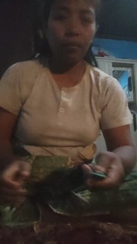 This media file is a short video clip showing a person in a dimly lit room. The subject is an individual, likely female, with dark hair and wearing a light-colored t-shirt with two buttons at the neckline. They are seated, and their hands are prominently featured as they appear to be engaged in some activity.

The person is holding a green object, possibly a vegetable or plant, and seems to be manipulating it with their fingers. The lighting is low, making it difficult to discern many details about the surroundings. However, a piece of patterned fabric, possibly a sarong or skirt, is visible draped around the lower body of the person. In the background, there is a glimpse of what appears to be furniture, possibly cabinets or shelves, and a window with a blue light emanating from it, suggesting artificial illumination.

The overall mood is intimate and focused on the task at hand, with the dim lighting creating a sense of privacy or perhaps nighttime. There are no discernible signs of weather conditions. The location context provided as "Sakra, Indonesia" suggests a potential cultural setting. No visible text is present in the video. The actions of the person, focused on the green object, imply a domestic or preparatory activity, such as cooking or crafting.