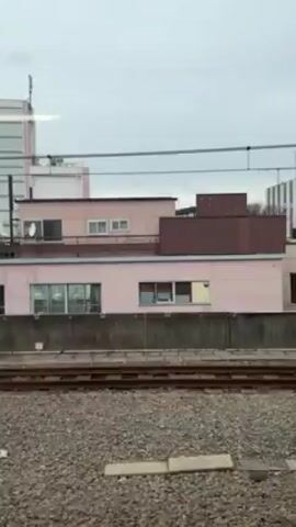From the perspective of a moving train, the landscape outside the window consists of a series of low-rise buildings. The structures are varied in color, ranging from pink and beige to green. A prominent multi-story building with a modern, striped design is visible in the background. Concrete barriers line the bottom of the frame, while the train tracks run parallel to the view. Overhead wires are also visible. The sky is overcast.
