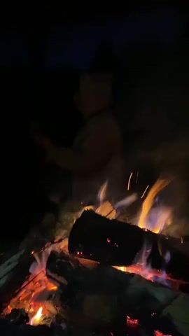 A person is standing in front of a bonfire at night, holding a phone. The bonfire is burning brightly with flames and embers. The background is dark, with trees visible in the distance. The person appears to be looking at the phone. The video is short and does not contain any dialogue.