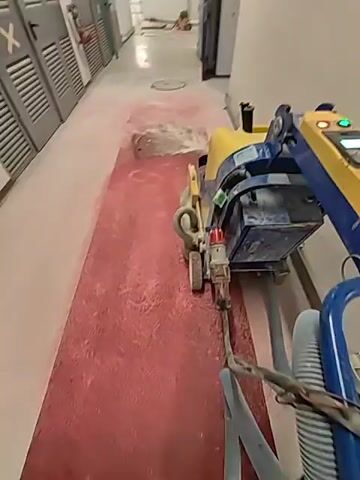 A floor grinding machine is being used to process a section of a hallway floor. The machine is yellow and blue and has wheels and a hose attached. The floor is covered in a pinkish dust from the grinding process. The hallway has doors on the left side and a white wall on the right. There are other pieces of equipment in the background.