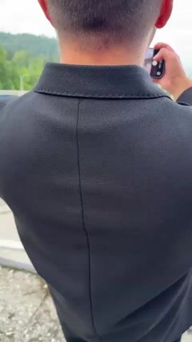 The video begins with a close-up shot of the back of a person's head and neck, wearing a black suit jacket. The person is holding a smartphone in their hands and appears to be taking a photo. The camera then pans out to reveal a wide shot of a large group of people posing for a photograph outdoors. The group, dressed in various formal and semi-formal attire, is arranged in front of a scenic background of rolling green hills, a body of water, and a cloudy sky. Many individuals in the group are raising their hands in the air and smiling. In the center of the group, a bride in a white wedding dress and a groom are visible. The person with the smartphone is positioned in the foreground, continuing to film the scene. The ground appears to be wet, suggesting recent rain. The overall atmosphere is celebratory.