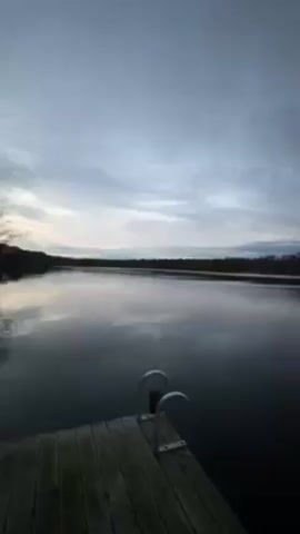 The video shows a calm lake under a cloudy, grey sky. A wooden dock with a metal ladder sits in the foreground. Trees line the opposite shore of the lake. The water reflects the sky, creating a mirrored effect. There are no people visible in the video. The camera is static, providing a wide shot of the scenery.