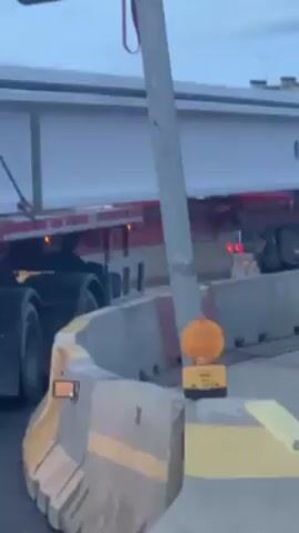 A large truck is transporting a wide, white beam through a street construction zone. The truck, pulling a flatbed trailer, is white with a red cab and displays the name "Despalles." The beam extends beyond the trailer's width, requiring careful maneuvering around concrete barriers painted with yellow stripes. A construction worker in a high-visibility yellow vest stands near the truck, guiding its movement. A triangular yield sign with a "100 m" distance indicator is visible in the foreground. The sky suggests it is either early morning or late evening. The construction zone is marked with orange warning lights and traffic control measures.