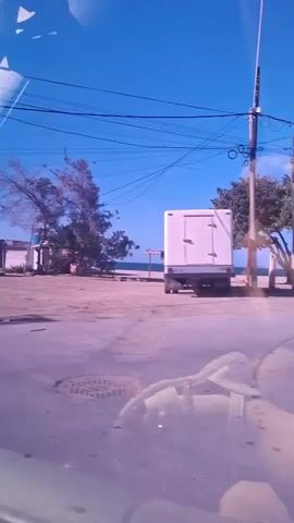The video shows a bright, sunny daytime scene from inside a moving vehicle, looking out the passenger window. The setting appears to be a coastal area, likely Playa Parguito, Venezuela.

In the foreground, an unpaved road with dirt and sand is visible. Reflections from the vehicle's interior, possibly the dashboard, are present on the window. Above, several power lines stretch across a clear blue sky.

The mid-ground features a mix of sparse vegetation and a few small, simple buildings or shacks. To the right at the beginning, a white truck is partially visible. A sign with the partially visible text "VENEZU" can be seen near the truck. As the vehicle moves, dense green trees become more prominent, lining the road. In the distance, beyond the trees, the sparkling sea is visible under the bright sky. People can be faintly seen gathered under the trees closer to the shore. The overall impression is a casual drive through a relatively undeveloped, sunny coastal landscape.