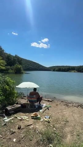 The video shows a wide shot of a lake on a clear, sunny day. A man sits in a striped beach chair under a white umbrella on the shore, facing the water. Fishing rods are set up near him. Further to the right, another man in a hat and shorts stands by his fishing gear. A third person, partially visible in the foreground, turns away from the camera, looking towards the lake. The shore is made of dirt and pebbles, with some scattered debris. The lake is surrounded by lush green trees on rolling hills under a bright blue sky with a few white clouds. The sun is visible in the upper left corner of the frame, casting a bright light.