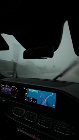 The video shows the interior of a car driving through heavy rain. The windshield wipers are active, clearing water from the view. The car's navigation screen displays a map. The weather appears overcast and foggy, reducing visibility. The passenger-side mirror and window are also visible, with raindrops accumulating on the glass. The interior lights of the car are illuminated, and a red light is visible on the lower right. The scene suggests the car is on a highway or a similar road with guardrails visible on the side.