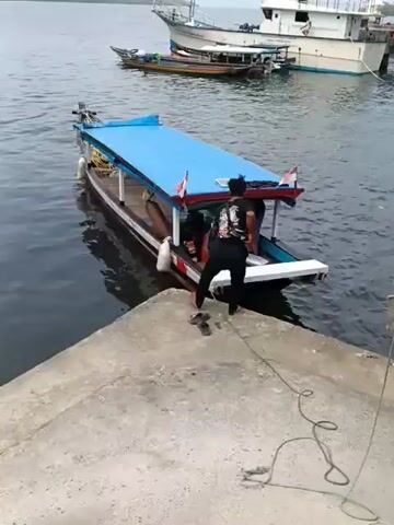 A person in a black t-shirt and black pants steps from a concrete pier onto a small boat with a blue canopy. Another person wearing a white cap and dark clothing is already on the boat. The boat has two Indonesian flags at the rear. In the background, several other boats are visible on the water, including a larger white vessel. The water appears calm and the sky is overcast.