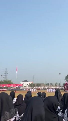 The video shows a wide outdoor shot of a ceremony or event on a bright, clear day. In the foreground, a crowd of people wearing black hijabs are partially visible from behind, their heads and shoulders filling the bottom of the frame. They appear to be spectators. In the middle ground, on a grassy field, a group of individuals dressed in white uniforms are performing a synchronized routine or dance. They are facing a line of other people dressed in formal attire, some of whom are wearing red sashes. A flag pole with the Indonesian flag (red and white) is visible to the left, with the flag slightly blurred by movement. In the background, there are buildings, utility towers, and trees. The overall atmosphere seems to be one of an organized public gathering.