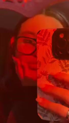 A woman with glasses and dark hair holds up a smartphone in front of a heart-shaped mirror with a pink frame, taking a selfie. The phone case has a floral pattern. The room is dimly lit with a red light casting a warm glow on the woman and her reflection. The woman’s hand and part of her face are visible in the mirror. She is wearing a ring on her finger. Music plays in the background.