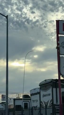 The video shows a street scene with a cloudy sky. There are several RVs parked behind a fence on the right side of the road. A traffic light is visible, and it is green. A street sign that says "Lexington St 2500" is hanging above the road. A white pickup truck is driving on the road. There is also a bike lane painted on the side of the road. The overall impression is that of a typical suburban street, possibly in the early morning or late afternoon given the lighting conditions.