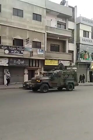 A video depicts a camouflage-painted armored vehicle driving on a street in a Middle Eastern urban area. The buildings lining the street appear to be low to mid-rise, with commercial signage in Arabic. There are pedestrians on the sidewalks and parked cars alongside the road. The vehicle has a turret-like structure on top and is moving slowly.