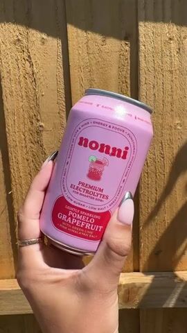 A hand holds a pink aluminum can of "nonni" brand electrolyte drink. The can has red lettering and graphics. The background is a wooden fence. The hand has manicured nails with a silver tip. The can is being rotated to show different sides.