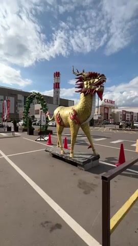 The video opens with a full shot of a large, golden dragon-like statue with red accents, standing on a stone platform in what appears to be a parking lot. The statue is facing the camera and is the main focus. The background shows a modern building with large windows and a red and white smokestack. The sky is cloudy with patches of blue. Orange traffic cones are visible around the statue. The camera slowly pans to the left, revealing more of the parking lot with parked cars and other decorative elements, including a green archway. People are seen sitting on a bench in the distance. The camera continues to move, showing the side of the statue and the surrounding environment. The sun is visible behind the clouds, casting shadows. The video ends with a shot of the statue from a slightly different angle, with the sky and buildings still in the background.