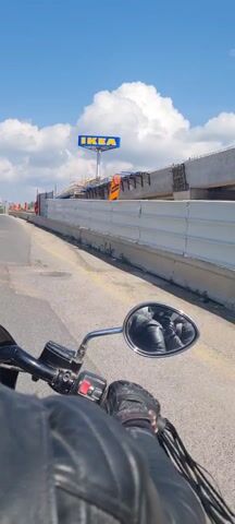 A person is riding a motorcycle on a paved road on a sunny day. The rider is wearing a black leather jacket and gloves. In the background, a large IKEA sign is visible, along with a bridge under construction. There are orange and black traffic markers along the roadside. The motorcycle's rearview mirror shows a reflection of the rider and the road behind. The sky is blue with white clouds.