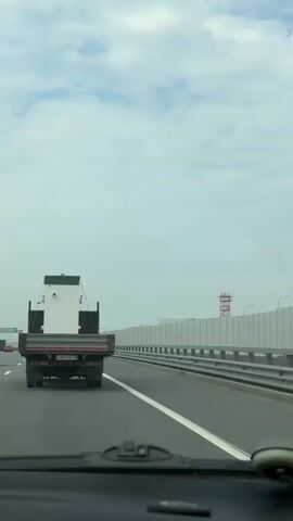 A car is driving on a multilane highway. Other cars and trucks are also traveling on the highway. The car with the camera is behind a flatbed truck carrying a large white object. The sky is cloudy. A tall tower is visible in the distance. Signage is visible above the lanes of traffic.
