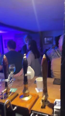 The video depicts a pub scene, taken from behind the bar. Several people are standing or sitting around the bar, seemingly enjoying themselves.  There is a barman (or woman) reaching up and adjusting something on the ceiling.  The lighting changes colors throughout the video. There are beer taps, glasses, cans of Coca-Cola, and other bar paraphernalia visible. The atmosphere is lively, possibly with music playing. Some people are clapping their hands.