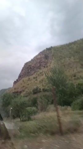 The video shows a rocky, vegetated mountainside under a cloudy sky. The camera appears to be moving, possibly from a vehicle, as indicated by the slight blur and the way the foreground elements shift. In the foreground, there are trees and some dry grass, along with a blurry wire fence. The mountain itself is steep, with a mix of exposed rock and green vegetation. The overall impression is of a natural, somewhat rugged landscape.