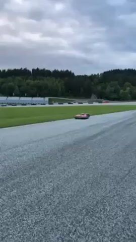 Here's a detailed description of the video:

**Scene:** A video shows an autonomous lawn mower moving along the edge of a racetrack.

**Environment:** The setting appears to be a racetrack or similar facility. There's a gray asphalt track bordering a wide strip of green grass. In the background, there are stands, trees, and a forest on a slight hill. The sky is overcast.

**Objects:**
*   Autonomous Lawn Mower: A rectangular, robotic lawn mower is the primary focus. It is black and pink, with wheels on either side.
*   Racetrack: The video captures a paved race track.
*   Grassy area: A wide patch of grass sits next to the racetrack.
*   Stands: There are bleachers or seating stands visible.

**Actions:**
*   The autonomous lawn mower is moving slowly along the edge of the racetrack and the grassy area. It appears to be cutting the grass or following a predetermined path.

**People:**
*   There is a figure visible at the edge of the frame on the left, seemingly observing the lawn mower.

**Overall Impression:** The video shows a robotic lawn mower performing its task near a racetrack, providing a practical and somewhat unusual juxtaposition.