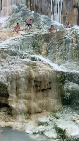 The video shows a natural landscape with people bathing in hot springs. The hot springs are surrounded by mineral deposits, creating a unique and beautiful environment. Several people are visible in the water, enjoying the therapeutic benefits of the springs. The water is a light blue color, and the surrounding rocks are covered in a white and green mineral buildup. The video captures the serene and relaxing atmosphere of the hot springs, highlighting the natural beauty of the location and the rejuvenating experience of soaking in the mineral-rich waters.