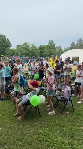 The video shows a crowd of people, mostly children, gathered outdoors on a grassy area with trees in the background. The sky is overcast. Many of the children are sitting on black folding chairs. Some children are standing, and some appear to be clapping or engaging in some kind of activity. There are several balloons visible, including green and yellow ones. In the background, there is playground equipment. Some adults are also present, some standing and some seated. The overall impression is that of an outdoor event or gathering for children.