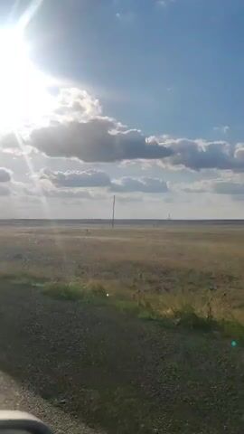 The video shows a view from inside a vehicle, looking out at a wide, open, grassy field under a bright, sunny sky with scattered clouds. The sun is prominently visible in the upper left quadrant, casting lens flares. A dirt or gravel road is visible in the foreground, with the side of the vehicle in the lower portion of the frame. In the distance, there are a few tall, thin poles, likely utility poles, and possibly some distant structures. The grass in the field appears dry and golden in some areas, with greener patches interspersed. The overall impression is of a vast, rural landscape.