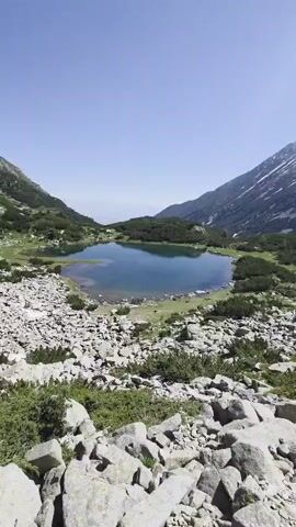 A handheld, vertical video captures a scenic view of a mountainous landscape with a clear blue sky. In the foreground, large, gray rocks and sparse green vegetation cover the ground. In the midground, a tranquil, dark blue lake is situated amidst rolling green hills dotted with trees. Further in the background, a majestic mountain range rises, with its peaks partially covered in snow and its slopes adorned with evergreen trees. The camera pans slightly, offering different perspectives of the lake and the surrounding natural beauty. At one point, the lower portion of the frame briefly shows a person's leg and knee, indicating the presence of a human observer.