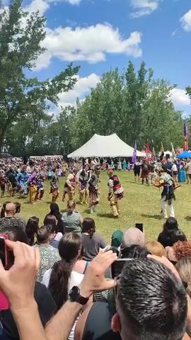 A crowd watches a group of people in traditional regalia dancing in a grassy field under a partly cloudy sky. The dancers are arranged in a circle and appear to be moving in unison. Several tents and trees are visible in the background. Many people in the foreground are holding up phones to record the event.