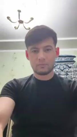 A man with dark hair and a beard, wearing a black t-shirt, is seated and looking directly at the camera. He makes a gesture, moving his hand across his face in front of his eyes.  The background shows a wall with a floral pattern, a stack of patterned cushions, and a ceiling fixture with multiple lights.