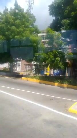 The video appears to be captured from a moving vehicle, possibly a bus or car, looking out a window. The scene shows a street with a paved road and a yellow curb. On the right side of the road, there are trees and buildings, including a tall, light-colored apartment building and a smaller building with an orange roof. Further in the background, a hillside is visible, covered with numerous small houses, suggesting a densely populated area. The sky is partly cloudy. In the foreground, a portion of the road surface and road markings are visible. There are no people clearly visible in the frame. The overall impression is of an urban or semi-urban environment on a sunny day.