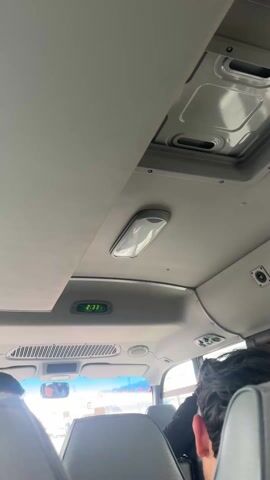 The camera, inside a vehicle, pointed towards the ceiling, shows two passengers with their backs turned to the camera. The ceiling contains a dome light, a clock reading 2:33, and sunroofs. Through the front windshield, stores are visible. Music plays.