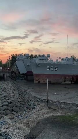 The video shows a low-angle shot of two large gray ships docked in what appears to be a dry dock or shallow body of water. The ship in the foreground has the number 925 painted in red on its hull. Both ships are gray with a red waterline. The sky above is a pale pink and orange, suggesting either sunrise or sunset. The ground around the ships is muddy and appears to be covered in debris, including what looks like rocks and hoses. There are also some wooden structures visible, possibly part of the dock. The overall scene suggests a static, possibly abandoned, environment.