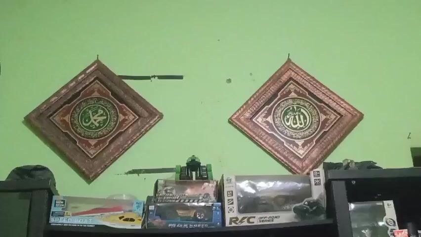 The video shows a wall with two framed Islamic calligraphy pieces hanging on it. The left frame contains Arabic script in green on a red and gold patterned background, enclosed by a square wooden frame. The right frame contains similar Arabic script in gold on a red and gold patterned background, also enclosed by a square wooden frame. Below the frames, on a shelf, are several toy boxes, including a remote control boat and remote control cars. A green toy robot or vehicle is positioned between the two calligraphy frames. The wall is painted a light green color.