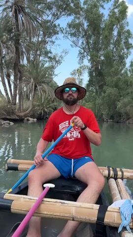 A man wearing a straw hat, sunglasses, a red t-shirt, and blue shorts is sitting in a canoe, paddling on a body of water. He is holding a blue and yellow paddle. The canoe has bamboo outriggers attached to its sides. In the background, there are palm trees, other trees, and a blue sky with some clouds. The water is calm and reflects the trees. There is another person in the canoe behind him, partially visible, holding a pink paddle. The man in the foreground is looking towards the camera.