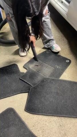 A person is vacuuming car floor mats in a garage, next to a white Volkswagen Polo. The person is wearing ripped jeans and colorful sneakers. The floor mats are black and covered in dirt and debris. The person is using a handheld vacuum cleaner with a brush attachment. The car is parked inside the garage, the door open. The garage has a metal roof and concrete floor.