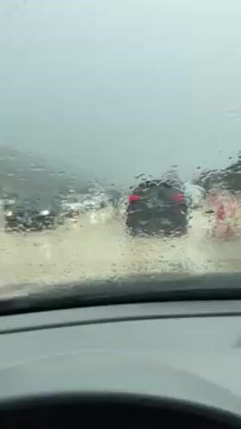 The video shows a flooded road from the perspective of inside a car. Raindrops cover the windshield as the wipers intermittently clear the view. Several cars are visible in front, navigating the flooded street. A person with an umbrella is walking in the water alongside the cars. The environment is overcast and gray, indicative of a rainy day. The car is stationary, suggesting it is part of the traffic stopped due to the flooding.