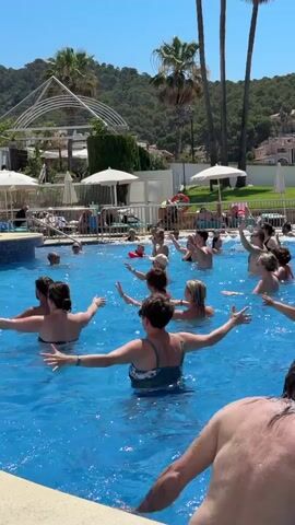 A group of people are participating in a water aerobics class in a bright blue swimming pool on a sunny day. The participants are in the water, some with their arms raised, others with their hands crossed over their chests. An instructor, wearing a white shirt and dark shorts, stands at the edge of the pool, facing the group and gesturing. In the background, there is a hotel building, palm trees, lush green hills, lounge chairs, and umbrellas. The water is clear and reflects the sunlight. Several other people are visible lounging by the pool or in the water.